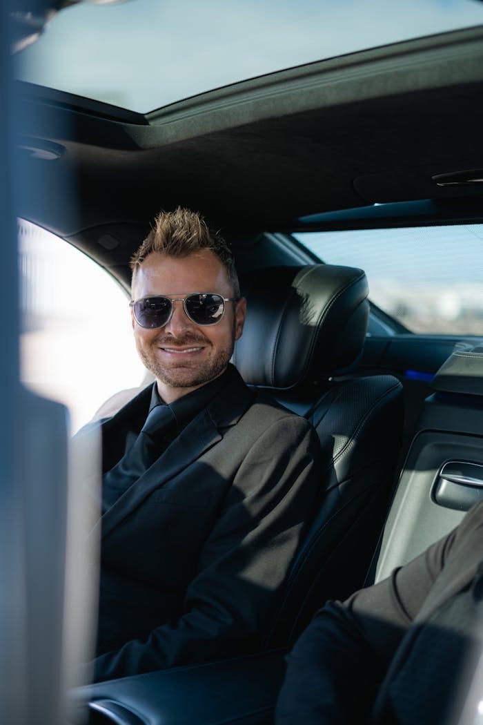 Smiling businessman wearing sunglasses seated in a luxury car interior, exuding confidence.
