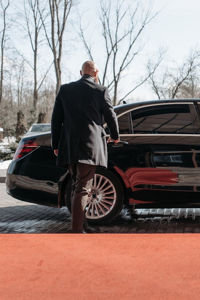 Bald man in a black suit approaches a luxury black car on a sunny day.